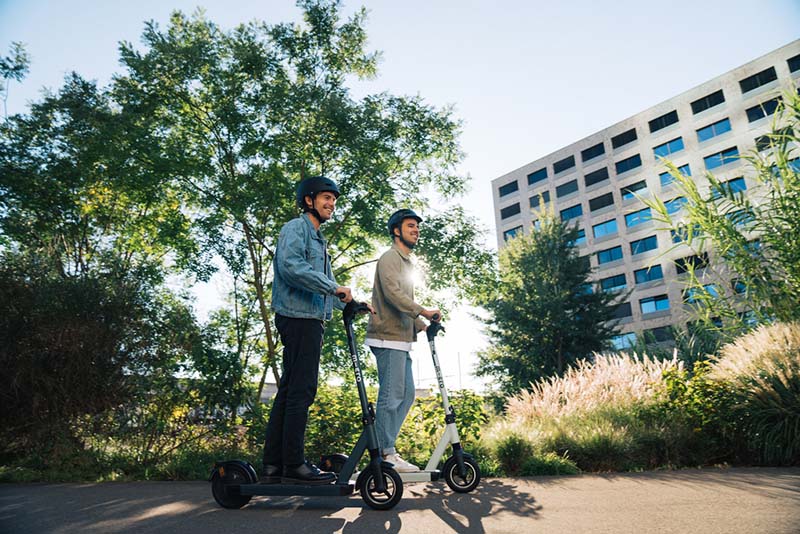 deux individus qui se déplacent en trottinette électrique dans un décor urbain et arboré